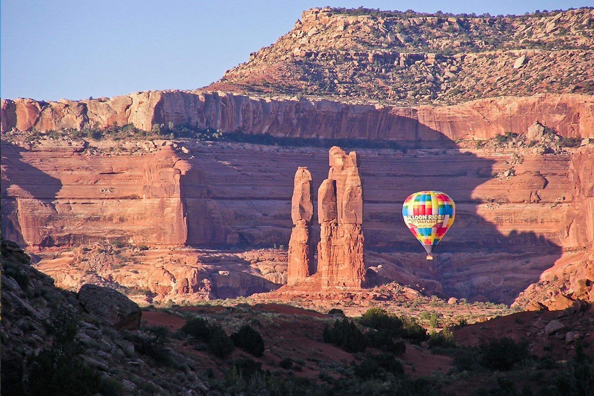 Hot air balloon floats past red rock formations in canyon.