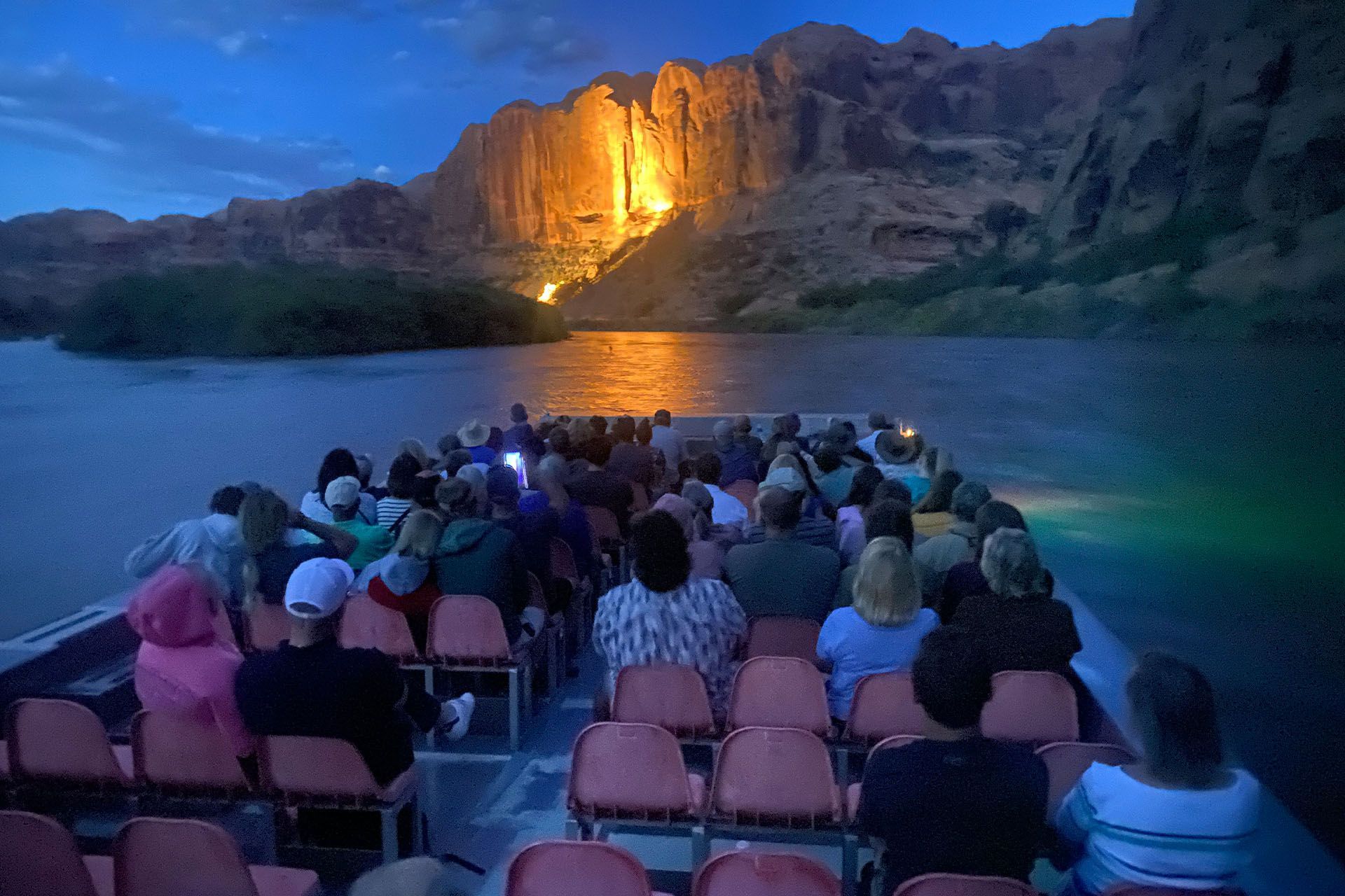 Boat tour on a river at dusk, illuminated rock wall in the background, passengers seated, various colors.