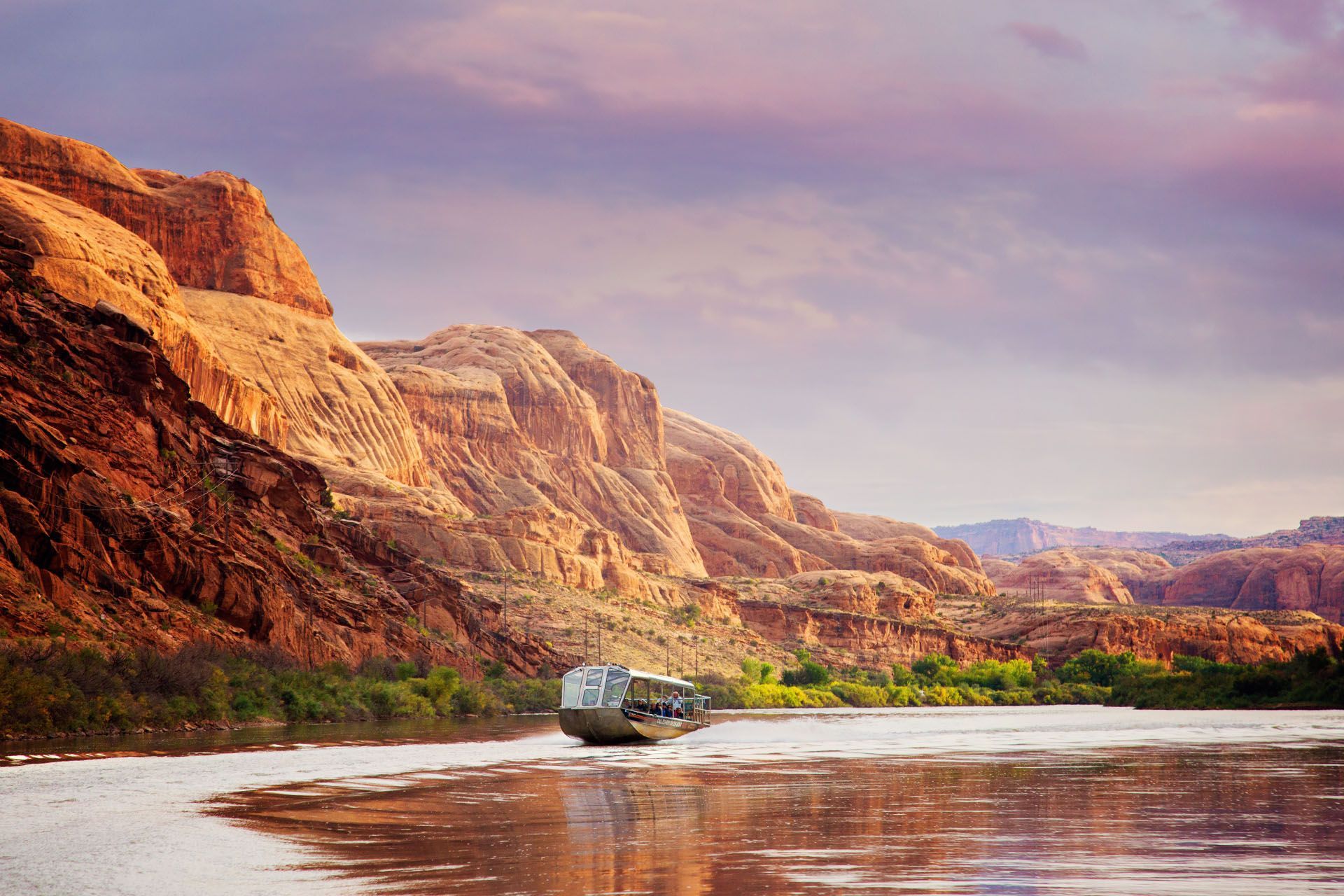 Boat on a river through canyons at sunset; orange rock formations, purple sky.