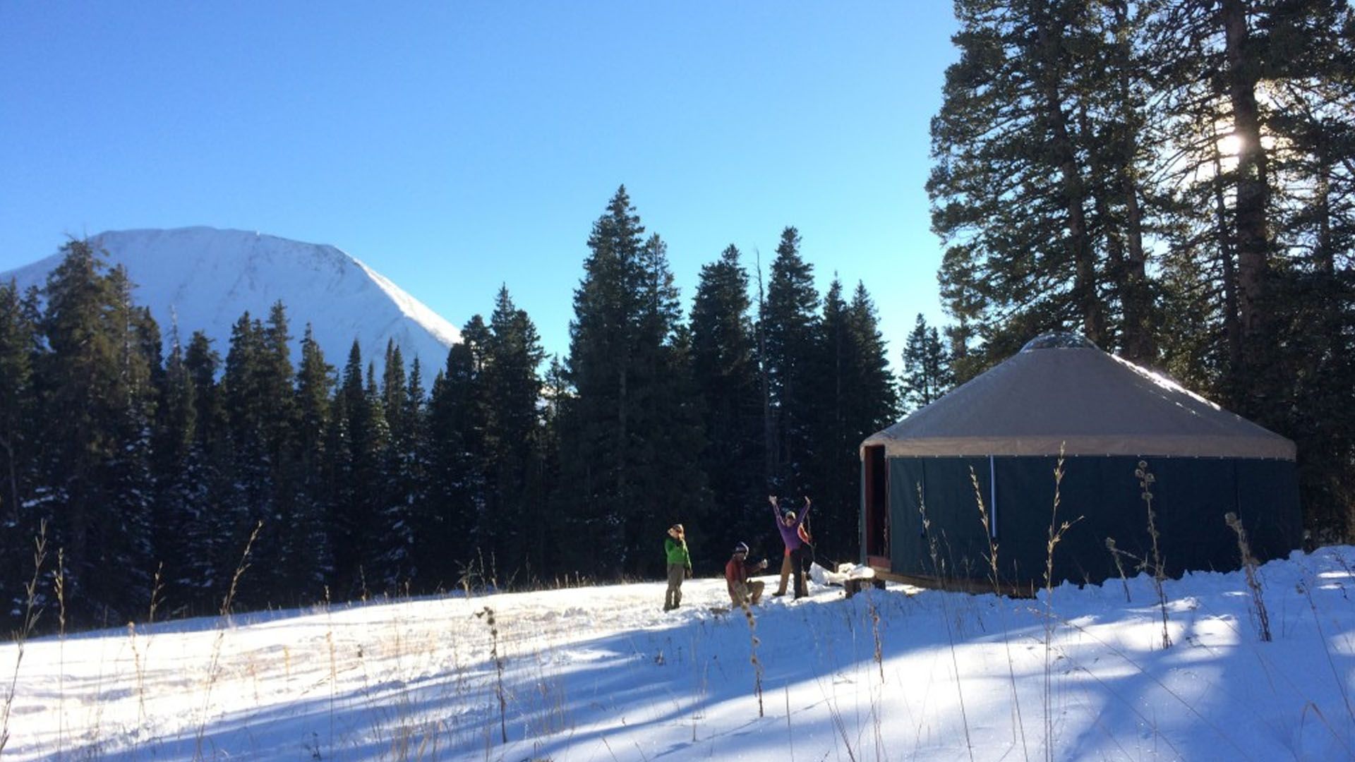 Snowy scene with a yurt, trees, and people. A mountain is in the background. Bright sunlight.
