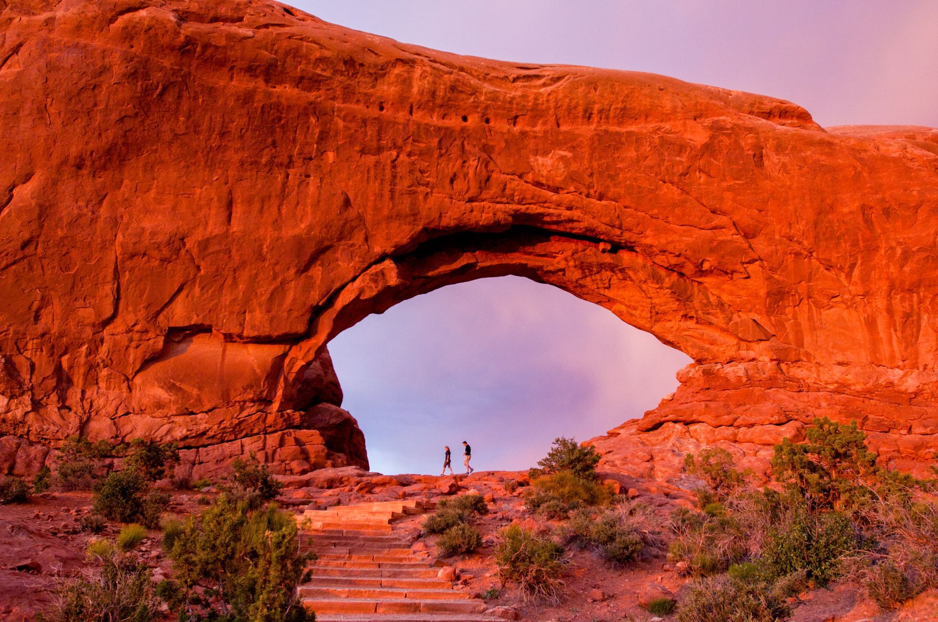 Red rock archway with two silhouetted figures on the horizon under a dusky sky.