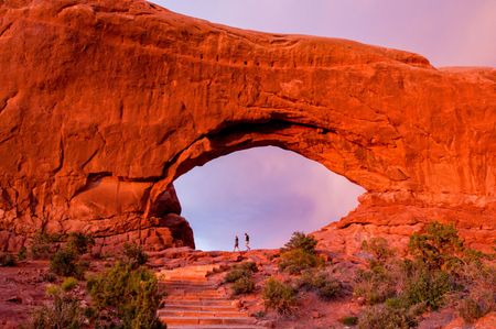 Red rock archway with two silhouetted figures on the horizon under a dusky sky.