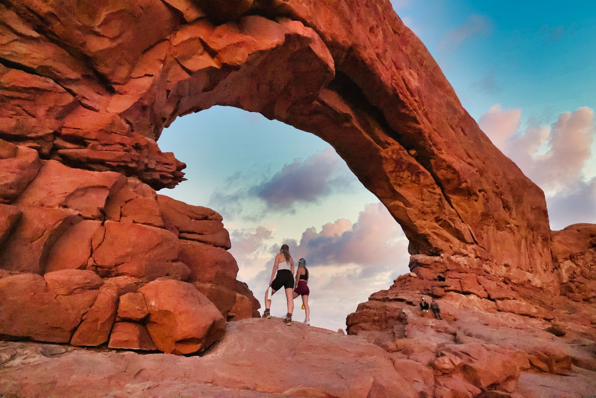 Two people stand under a large red rock arch against a sky of blue, pink, and clouds.
