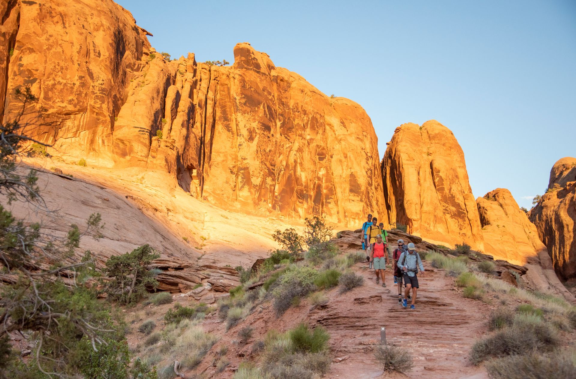 Hikers ascend a rocky desert trail toward tall, sunlit rock formations.