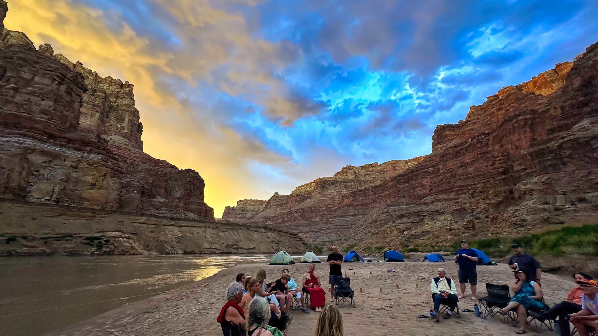 Campers gather on a sandy riverbank beneath canyon walls at sunset. Tents are pitched, sky is colorful.