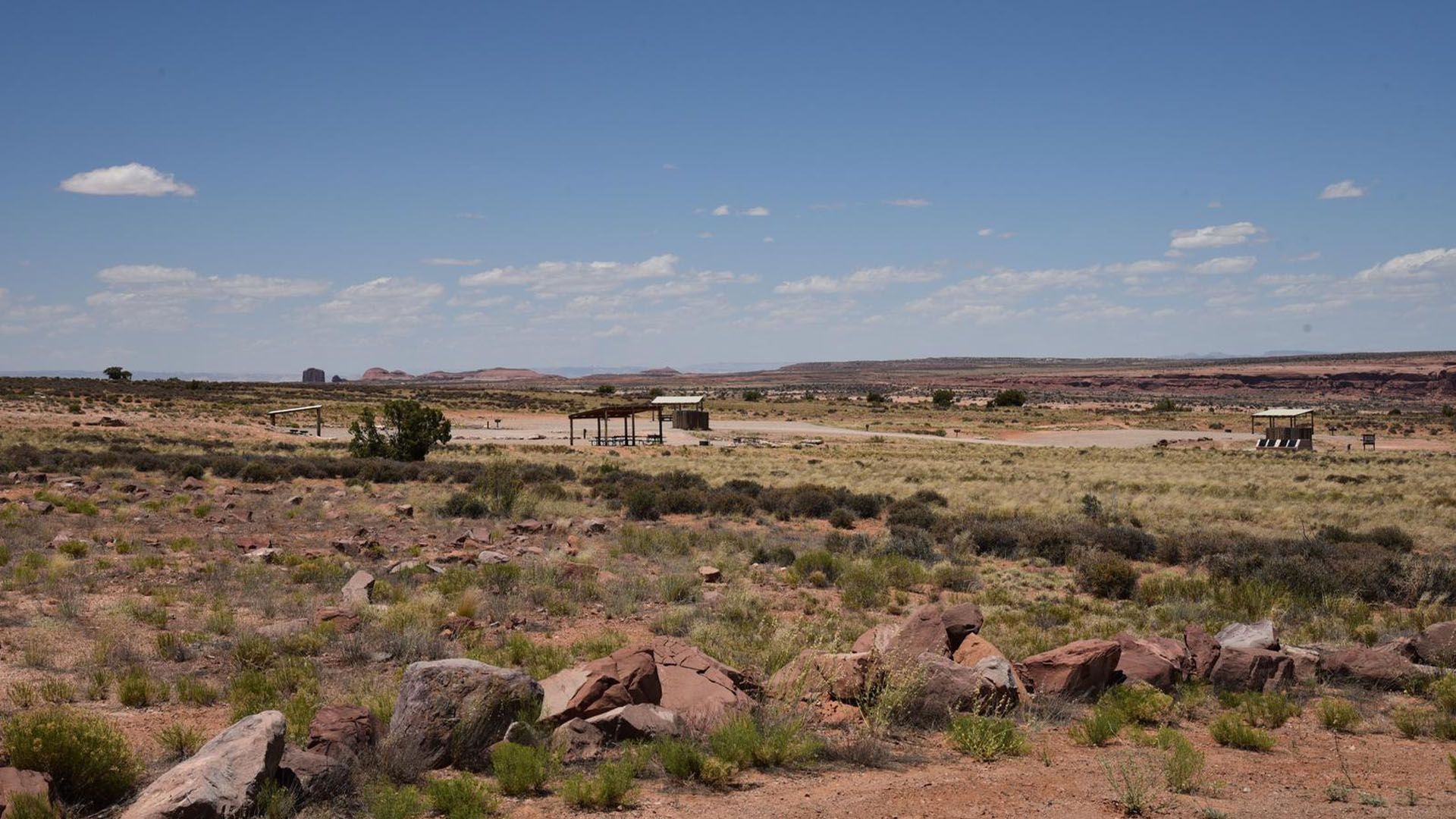 Desert landscape with sparse vegetation, brown earth, and blue sky; shelters are scattered in the distance.