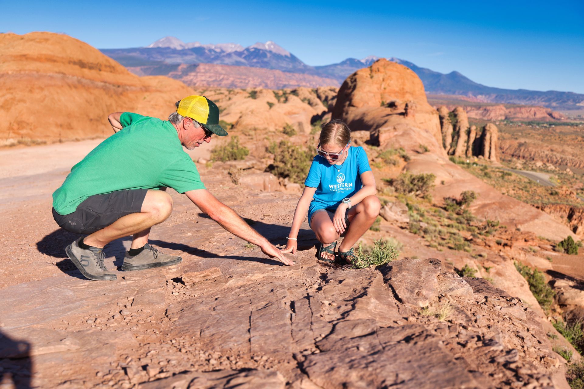 Hummer driver and guide explains dinosaur tracks to a guest as they squat down and look at the tracks in the rock.