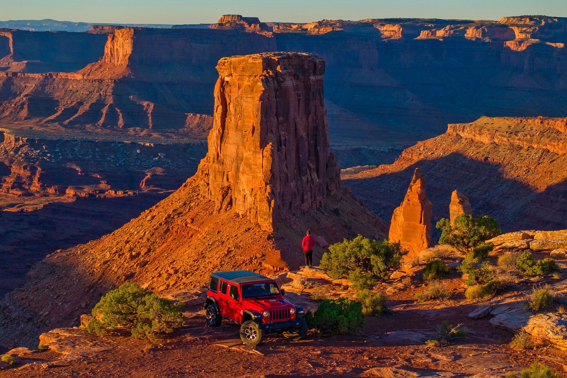 Red jeep parked near a large, sunlit rock formation in a desert canyon. Two people stand nearby.