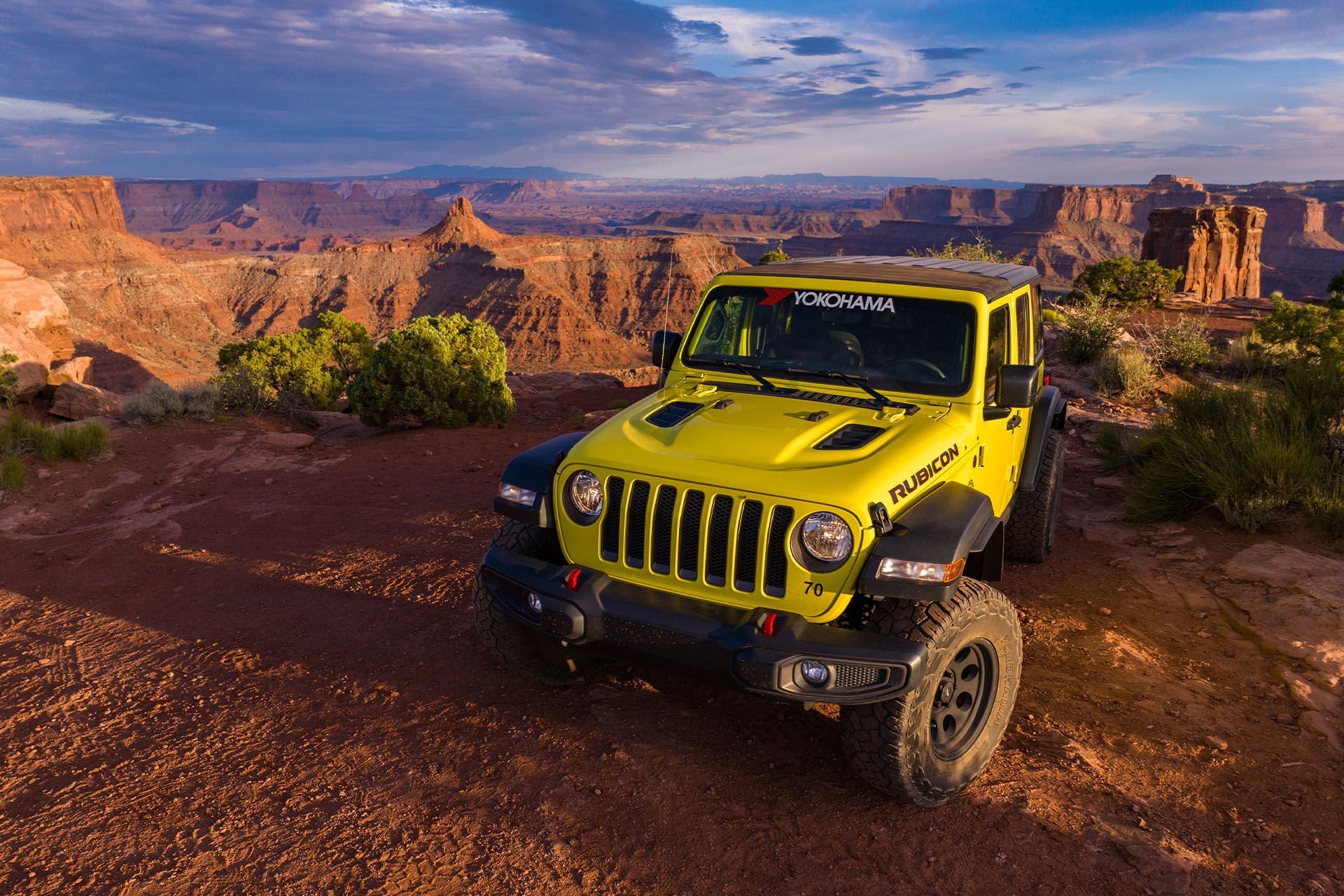 Yellow Jeep parked on a red rock cliff overlooking a canyon at sunset.