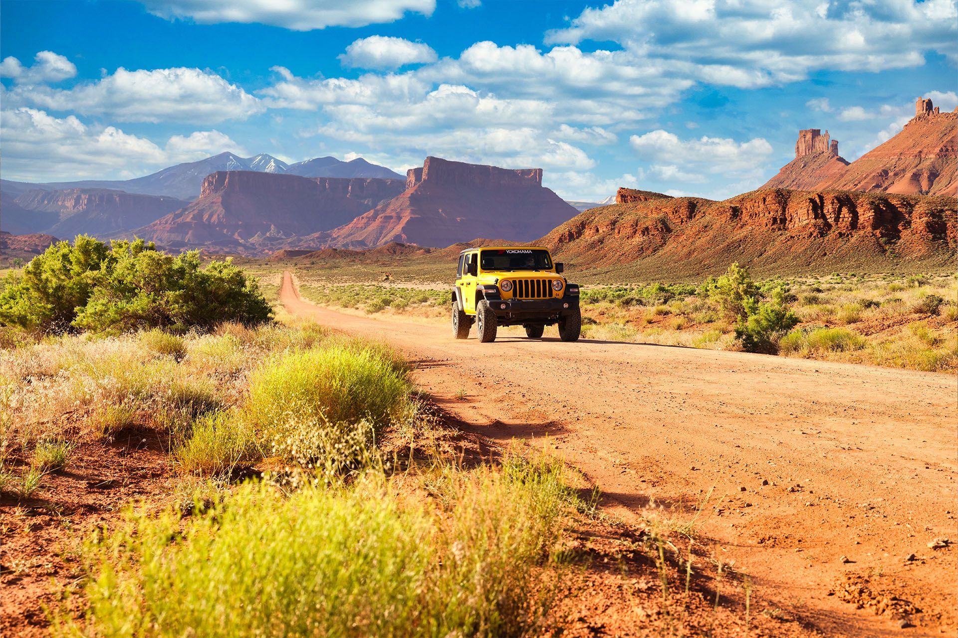 Yellow jeep driving on a dirt road through a desert landscape with red rock formations and blue sky.