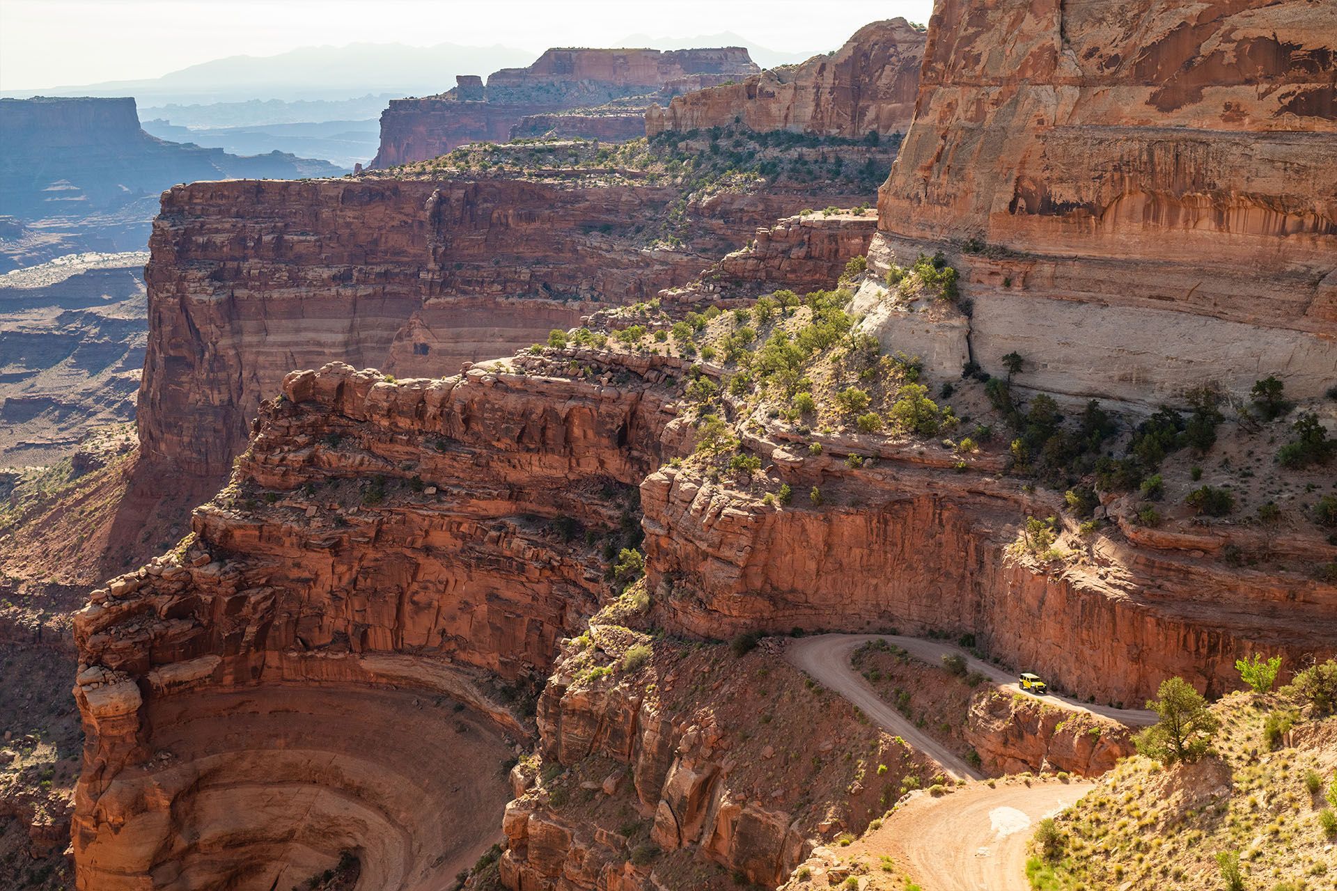 Red rock canyon landscape with a winding dirt road along the edge of a cliff, under a sunny sky.