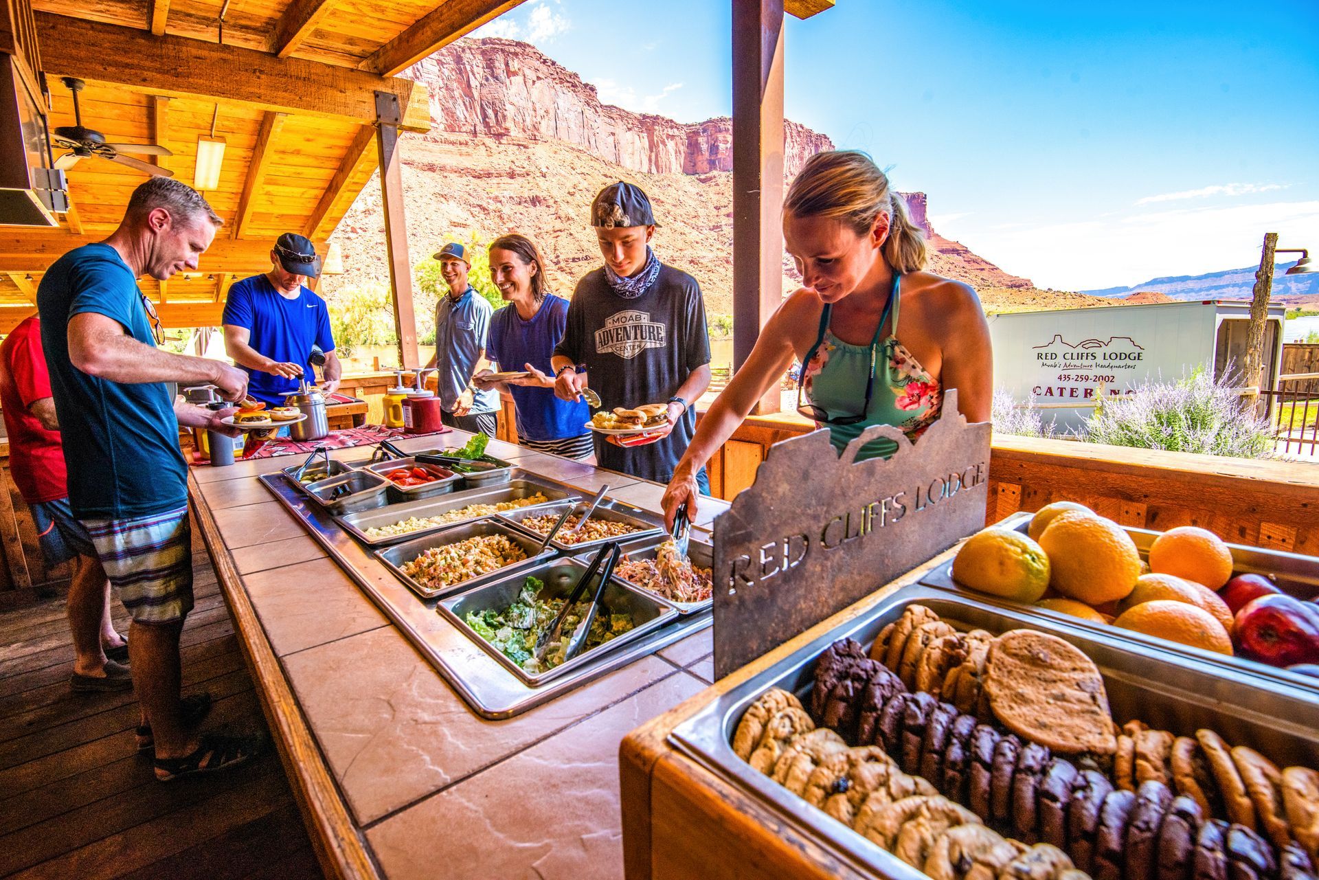 People serving themselves at a buffet, outdoors with red rock mountain backdrop. Cookies, fruit, and salads.