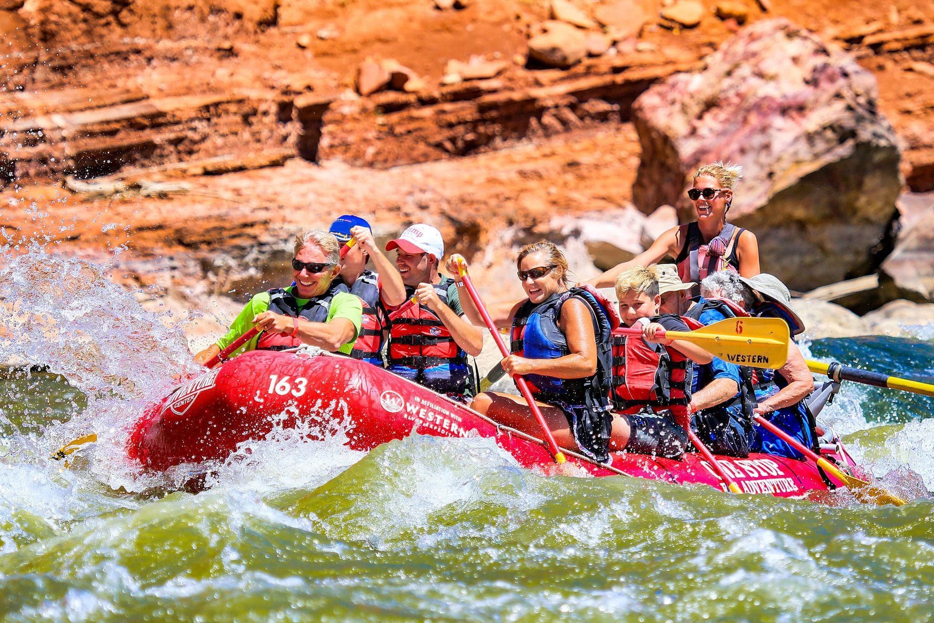 People white water rafting in a red boat on a river with rocky, brown canyon walls. Water splashes around them.