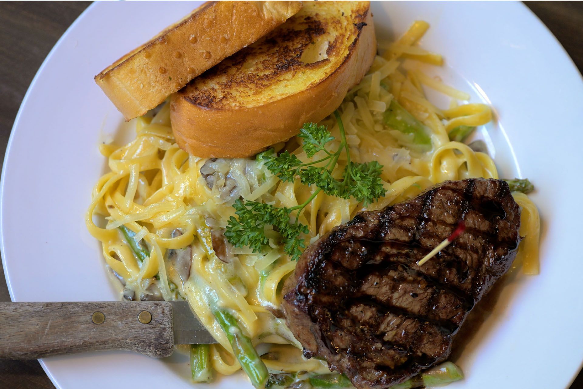 Steak, pasta Alfredo with asparagus, garlic bread, and knife on a white plate.