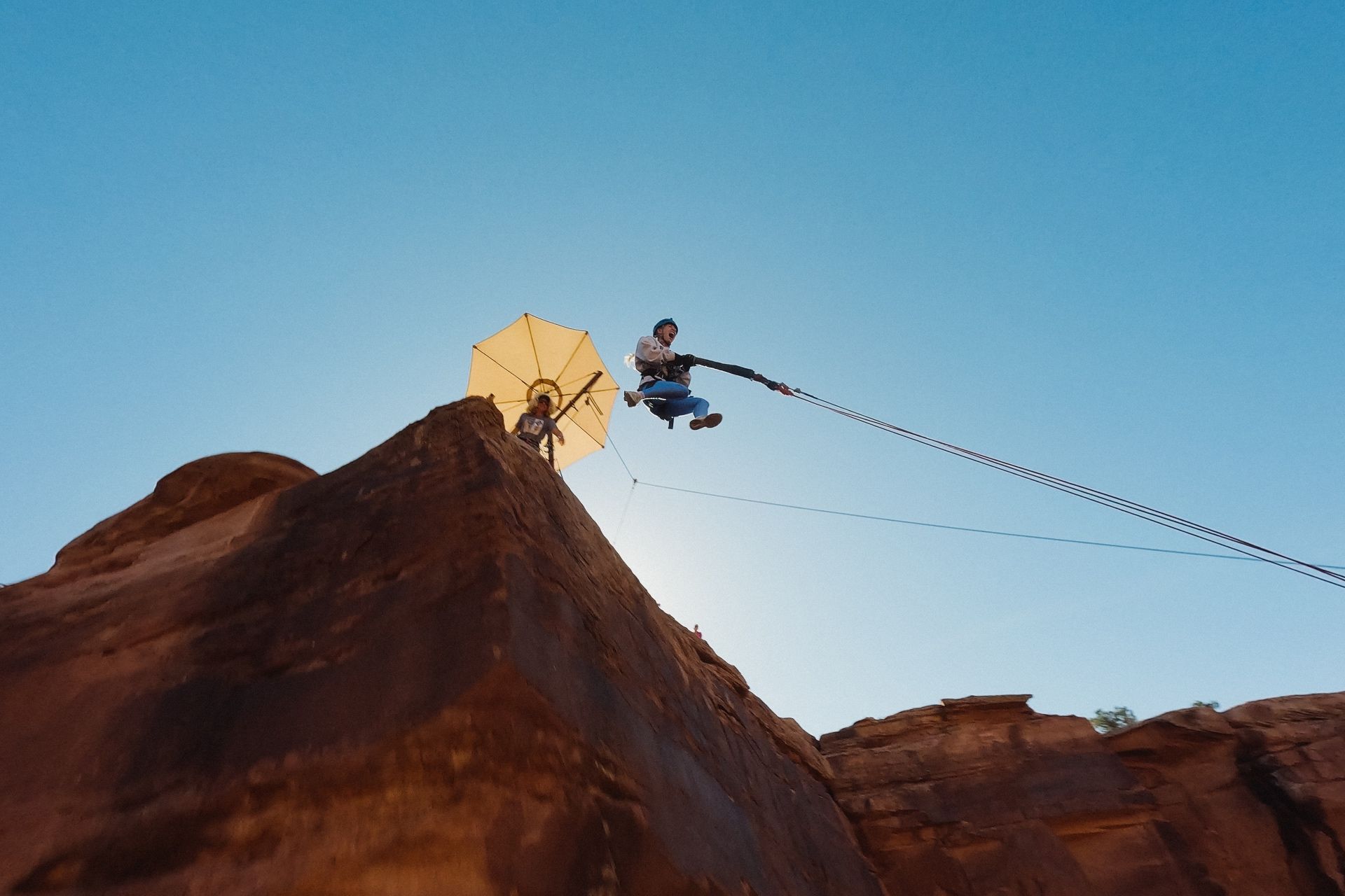 Person walks a suspension bridge over a desert canyon; red rock formations and blue sky.