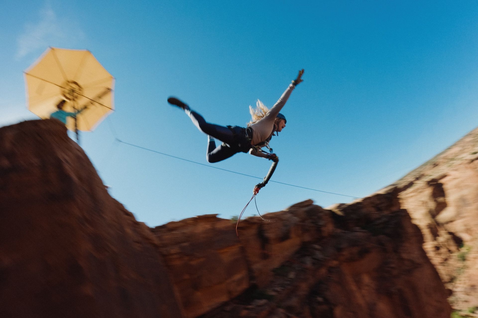 Person ziplining over a desert landscape, wearing a helmet and harness, against a blue sky.