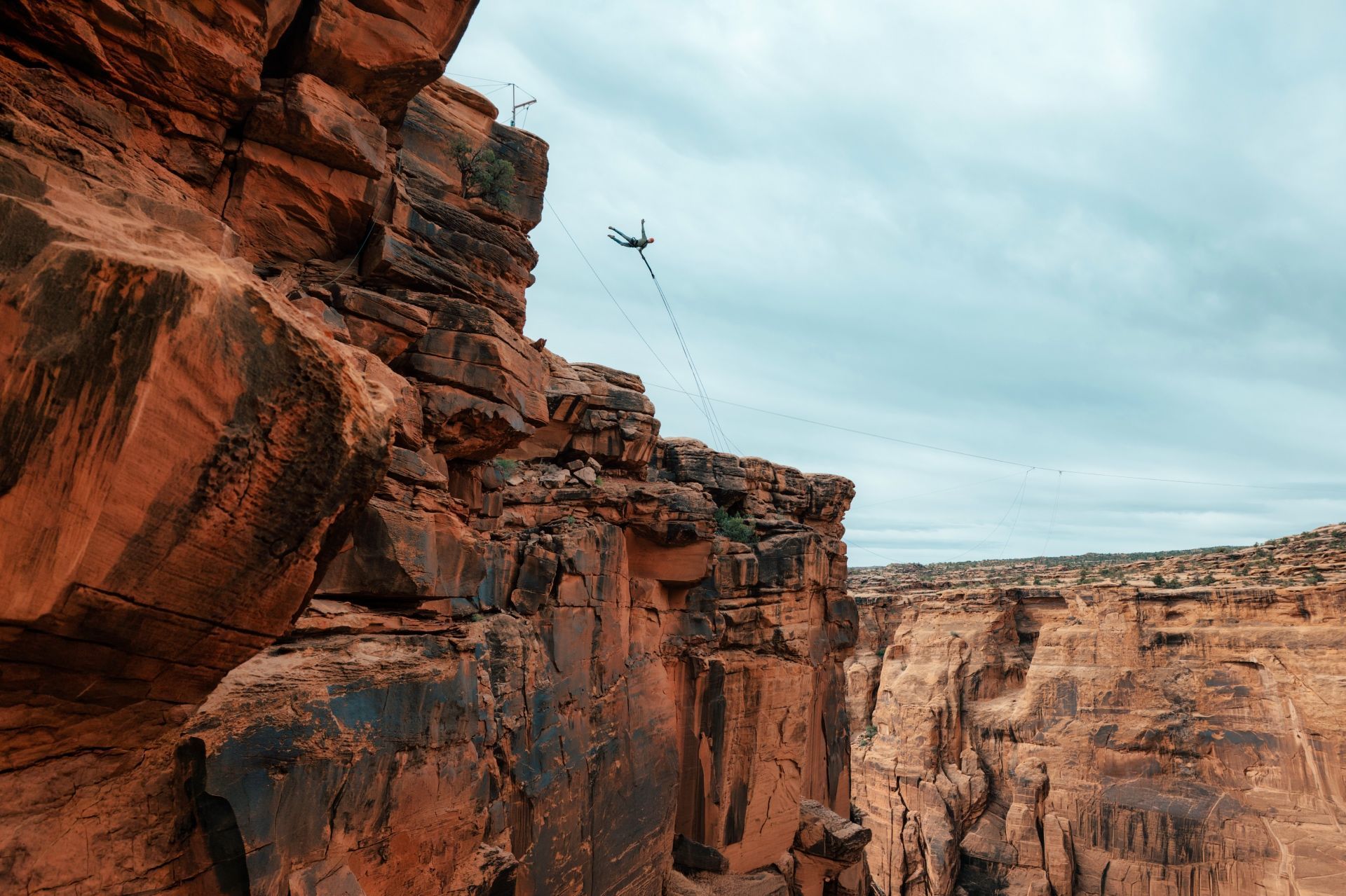 Red rock canyon cliffs beneath a pale blue sky, with a rope line spanning the gap and a person jumping off a cliff.