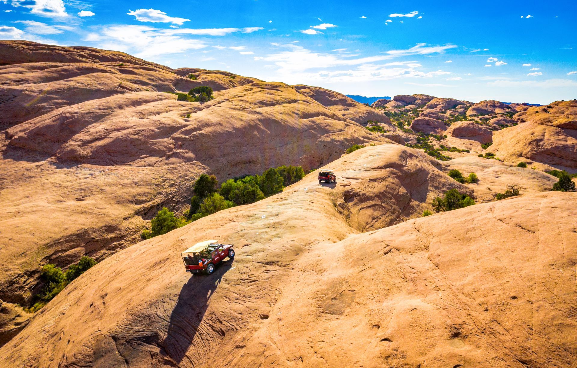 Two off-road Hummers driving across a sunlit Hells Revenge trail among rocky hills under a blue sky