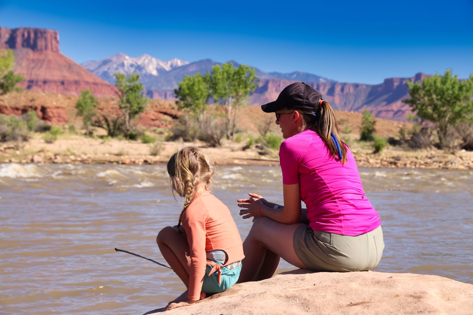 Woman and child sit on a rock by a river, mountains in the background.