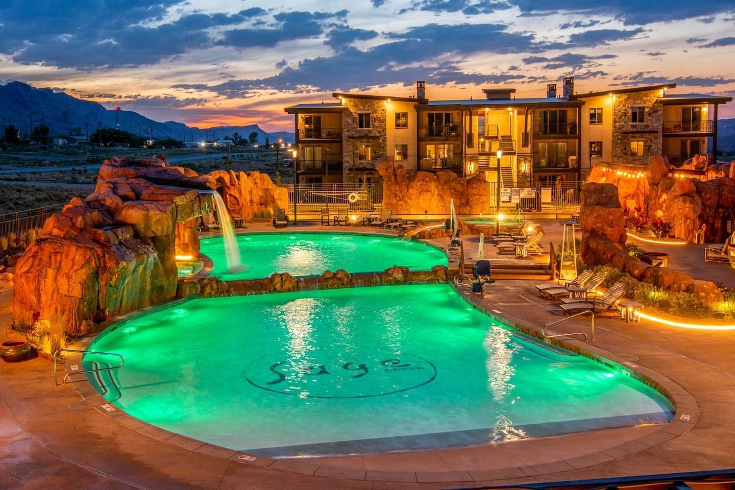 Sunset view of resort pools with a waterfall feature and lit-up buildings.