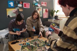 Three people play a board game on a table in a room with posters. Two look at the game, one holds cards.