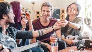Four friends smile and interact in a living room, holding drinks, a guitar, and a ukulele.