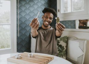 Man holding a pre-rolled cigarette and a bud in a kitchen, smiling with tongue out, table with a wooden board.