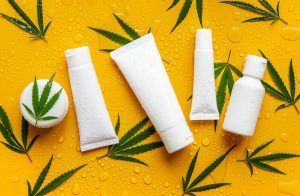 White cosmetic tubes and jar with green cannabis leaves on a yellow surface.