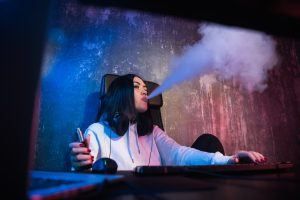 Woman exhaling vapor from a vape while using a computer in a room with blue and red lighting.