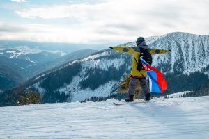Person on skis with arms outstretched, holding a red and blue flag, facing snow-covered mountains.