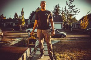 Man standing on a sidewalk holding a box, in front of houses and cars.