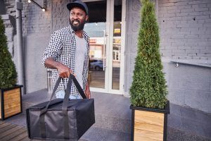 Person holding a black insulated bag exiting a building with plants on either side.
