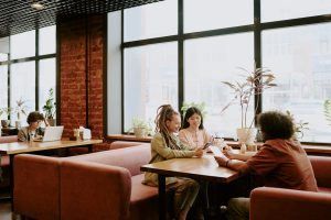 Three people seated around a table in a cafe, looking at a device. A person is working on a laptop at another table.