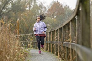 Woman running on wooden bridge, wearing jacket and leggings, fall setting.