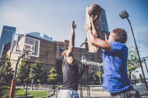 Two men playing basketball outdoors; one shoots, the other defends. City backdrop, sunny day.