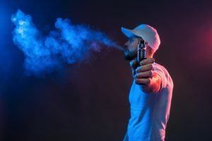 Person vaping, exhaling blue vapor; illuminated by blue and red light.