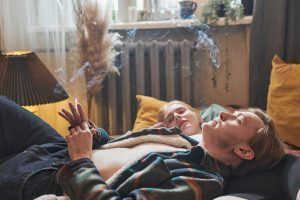 Two people relaxing on a couch, one holding a lit item. Smoke visible. Indoors.