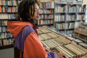 Person browsing records in a music store, wearing a purple and orange jacket, bookshelves in background.
