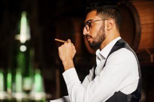 Man in glasses and vest holding cigar, looking thoughtful near barrels and bottles.