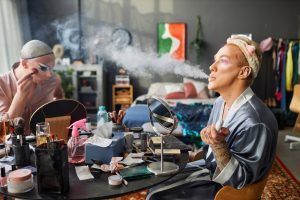 Two people applying makeup, one exhaling vapor. Inside a room with cluttered vanity.
