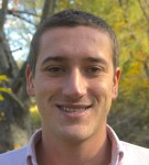Man with short brown hair smiling outdoors, wearing a light pink shirt. Trees in background.