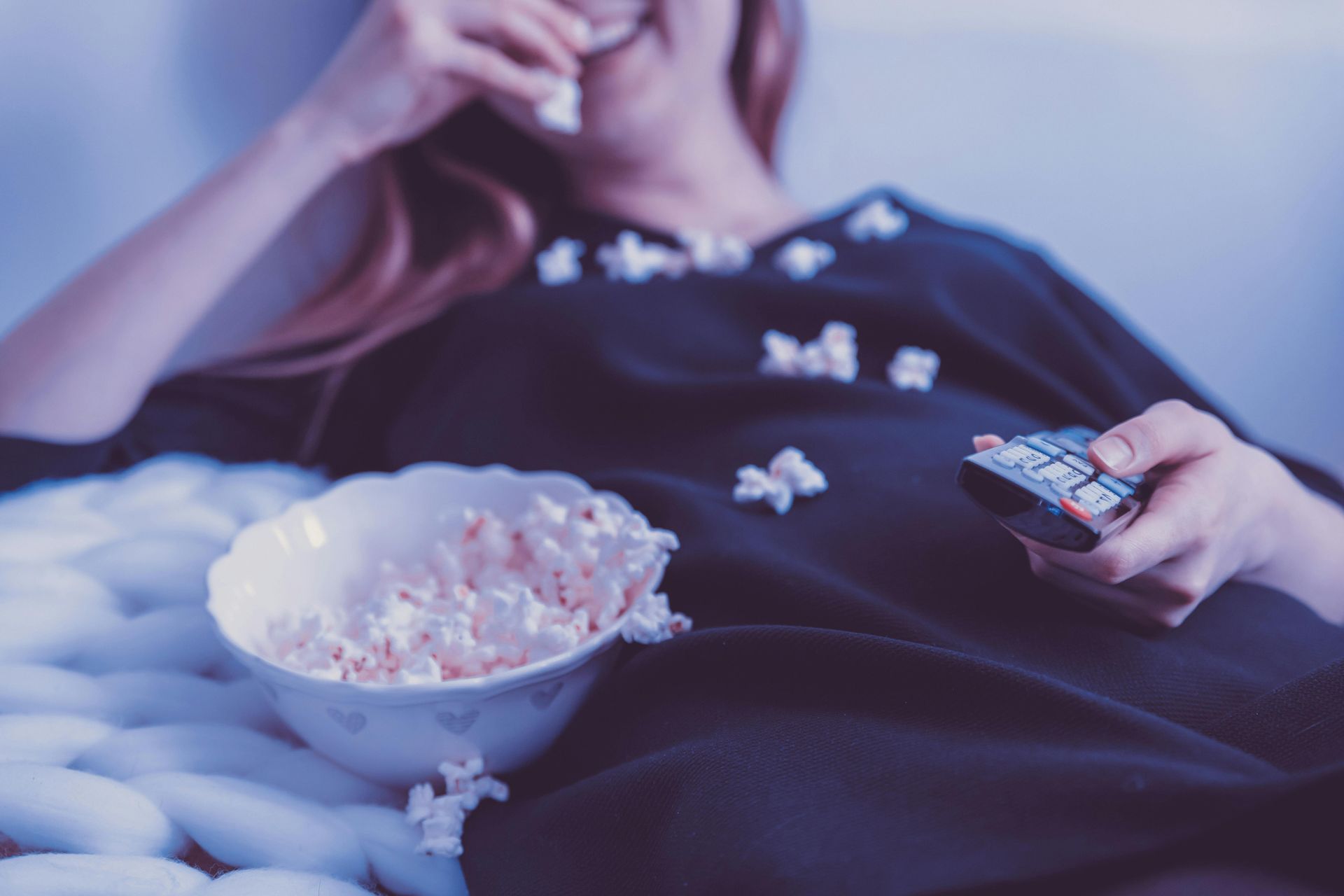Woman on couch eating popcorn and holding a remote, watching TV.