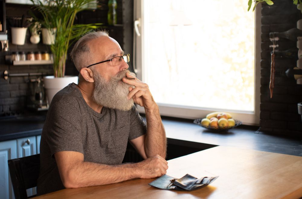 Man with glasses and a beard thoughtfully looks out a window in a kitchen, money on the table.