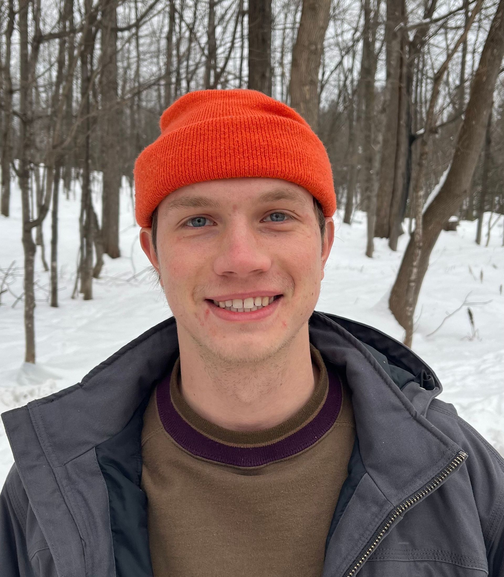 A young man wearing an orange hat and a jacket is smiling in the snow.