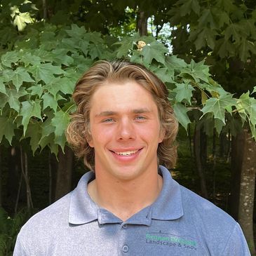 A young man in a blue polo shirt is smiling in front of trees.