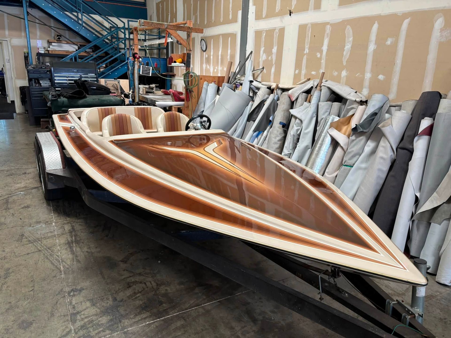 Wooden speedboat on a trailer in a workshop, with brown and white striped deck and stored materials nearby