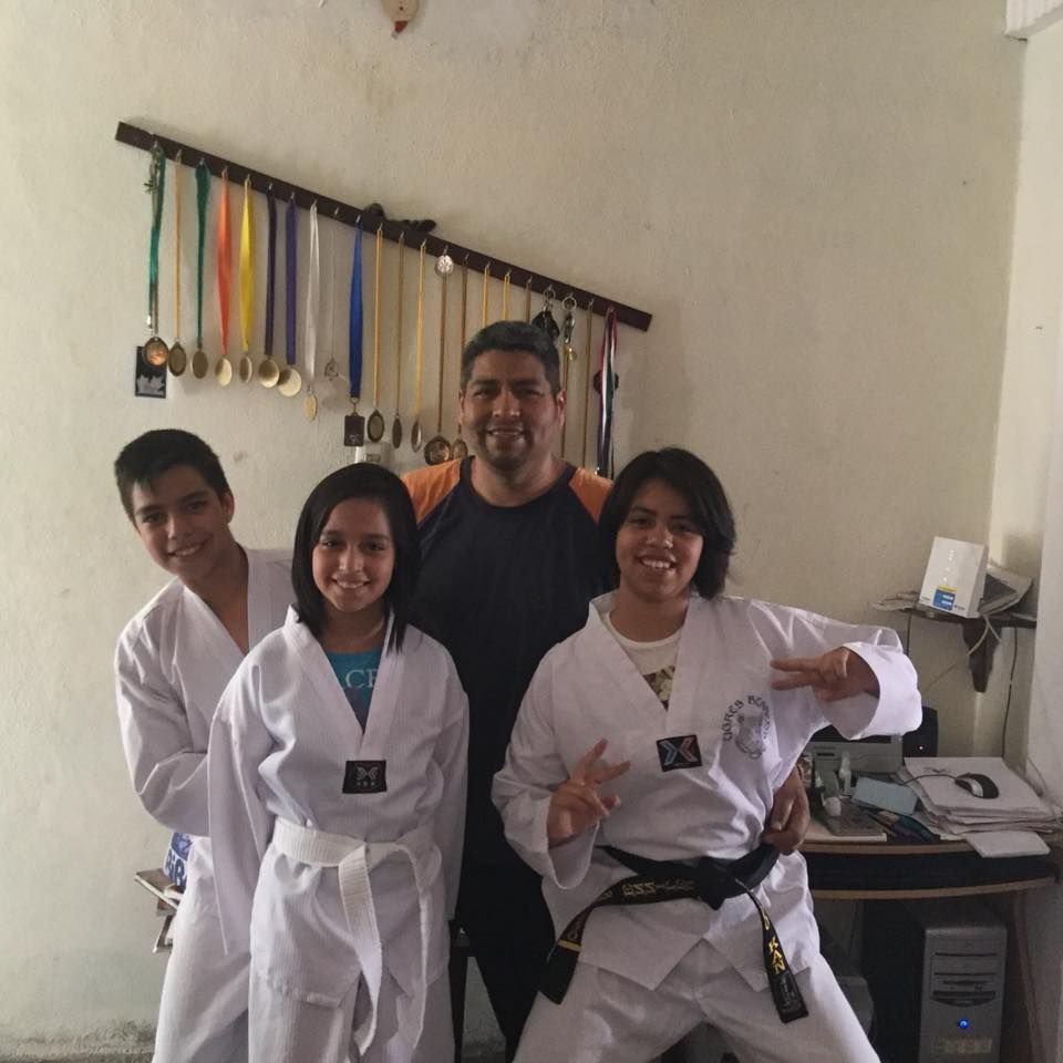 Four martial arts students in white uniforms and a coach pose indoors, smiling and flashing peace signs
