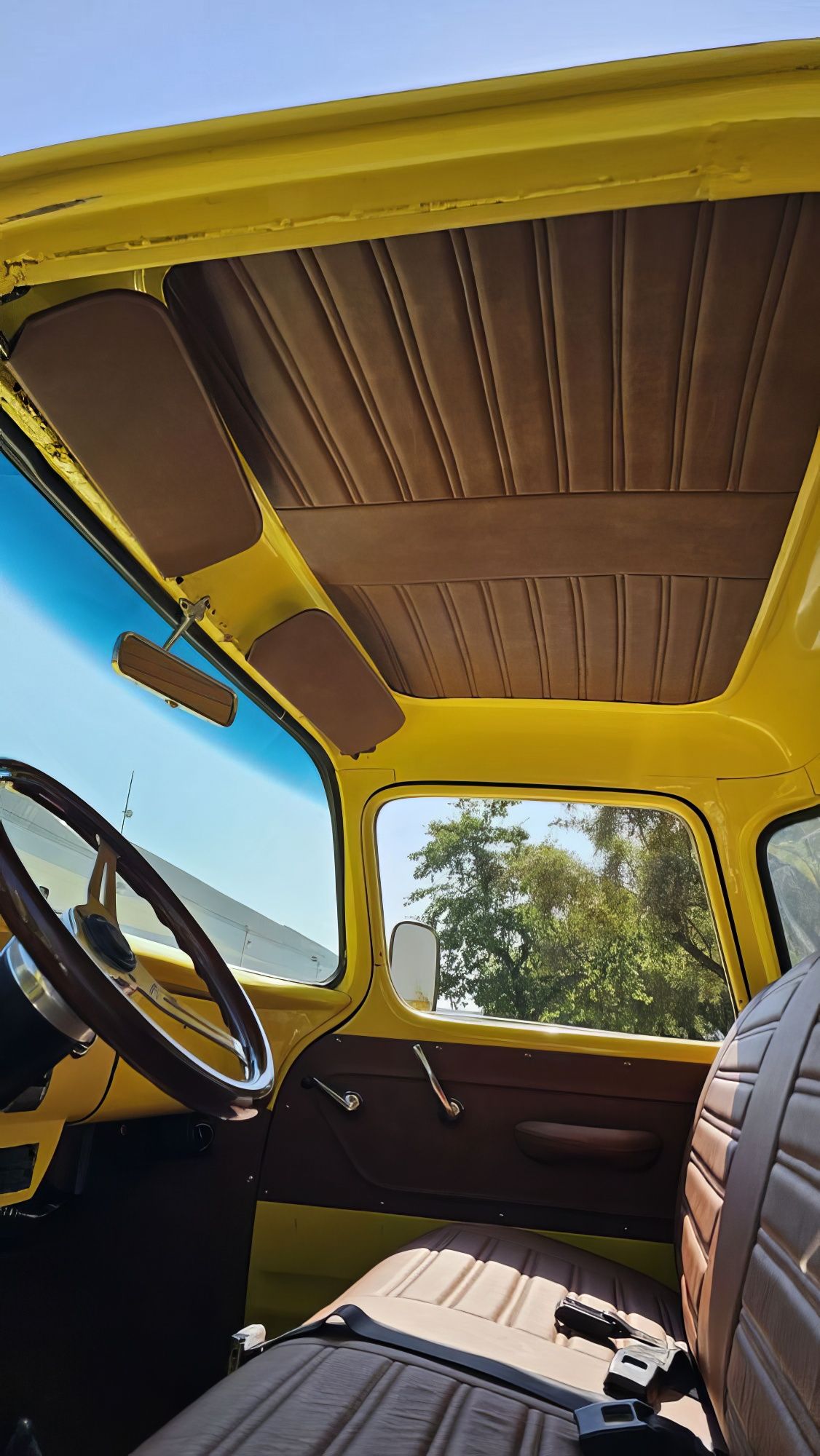Yellow car interior with steering wheel, brown seats, and an open roof showing blue sky and trees