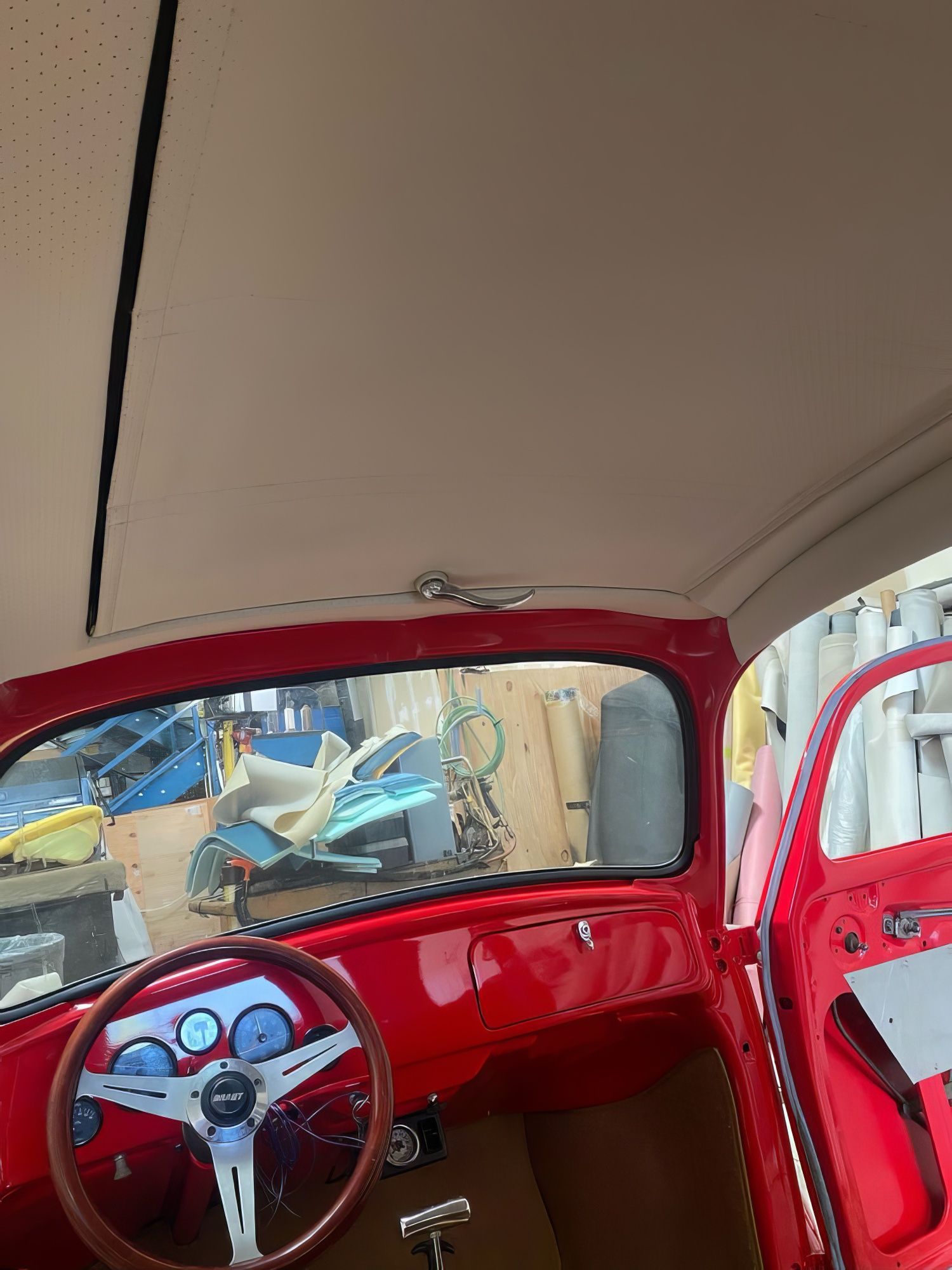 Interior of a red vintage car with a white roof, steering wheel, and a glimpse of tools outside the rear window