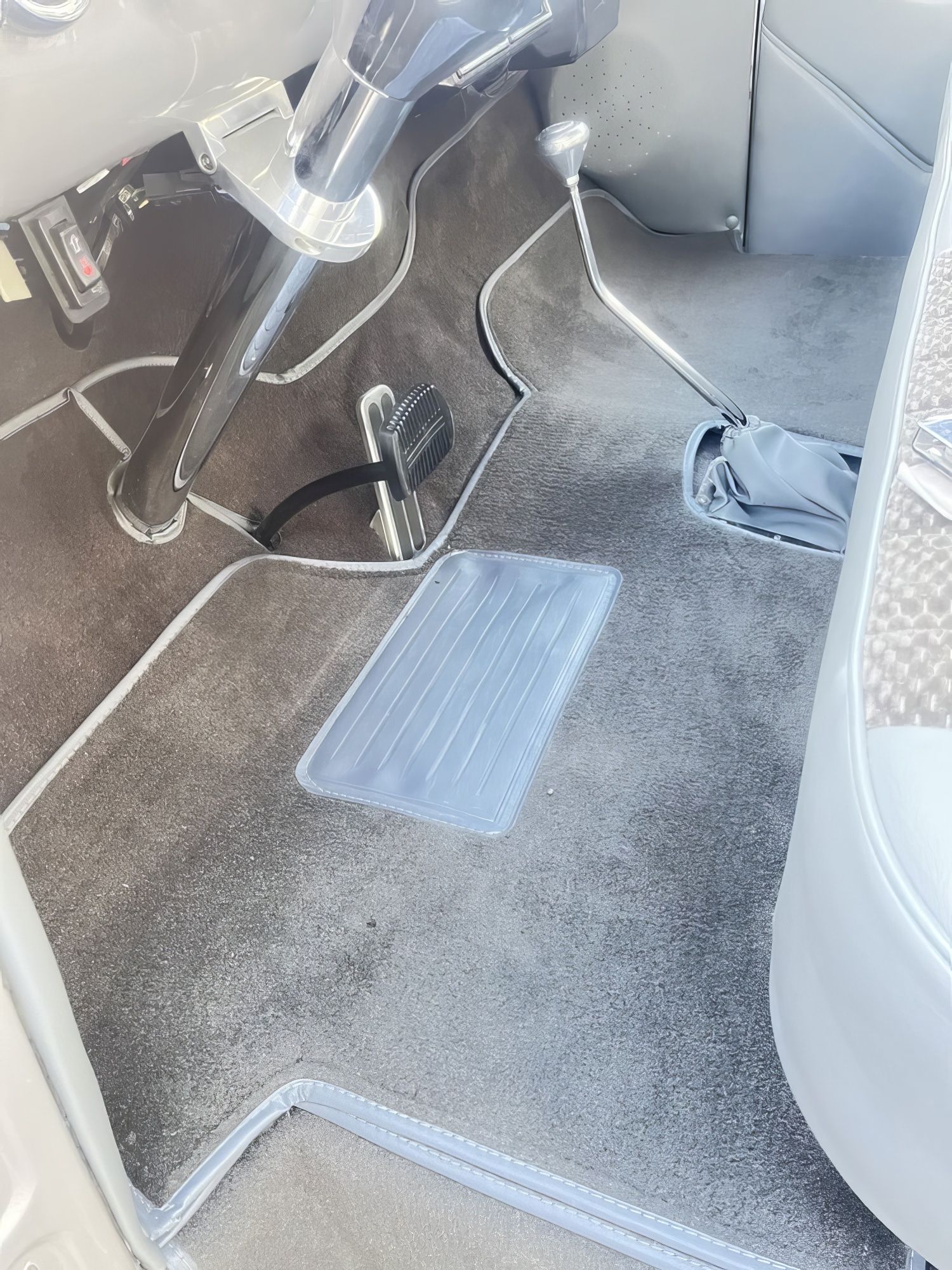 Car floor with gray carpet, pedals, and a clear plastic protective mat near the driver seat